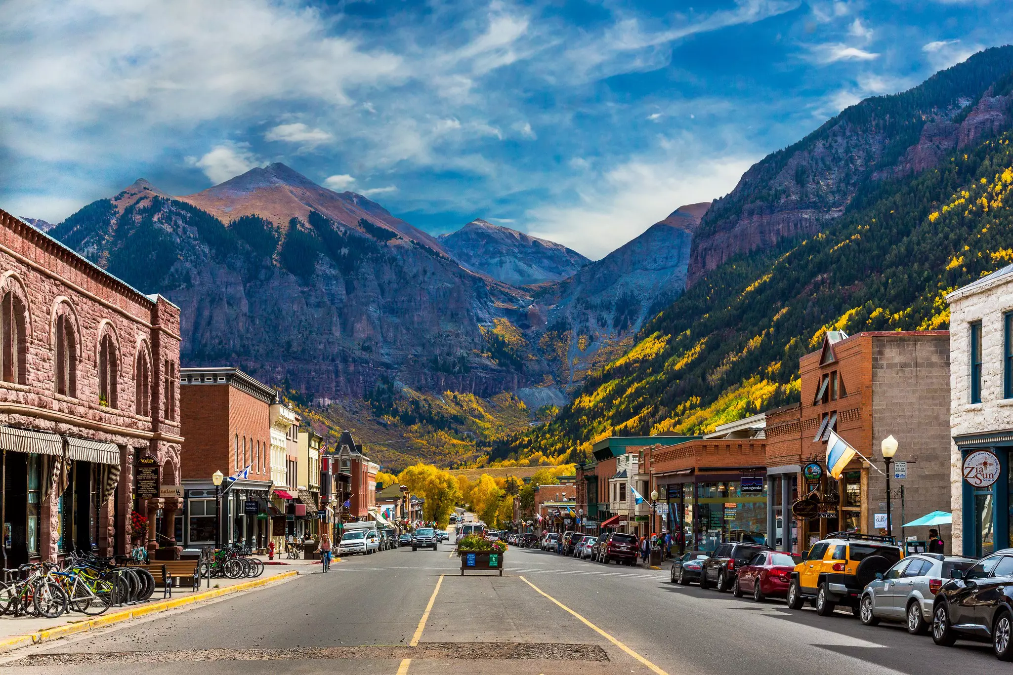 A street in a small town is lined with low buildings with shops. Mountains are seen in the distance, their slopes covered with trees in fall foliage.