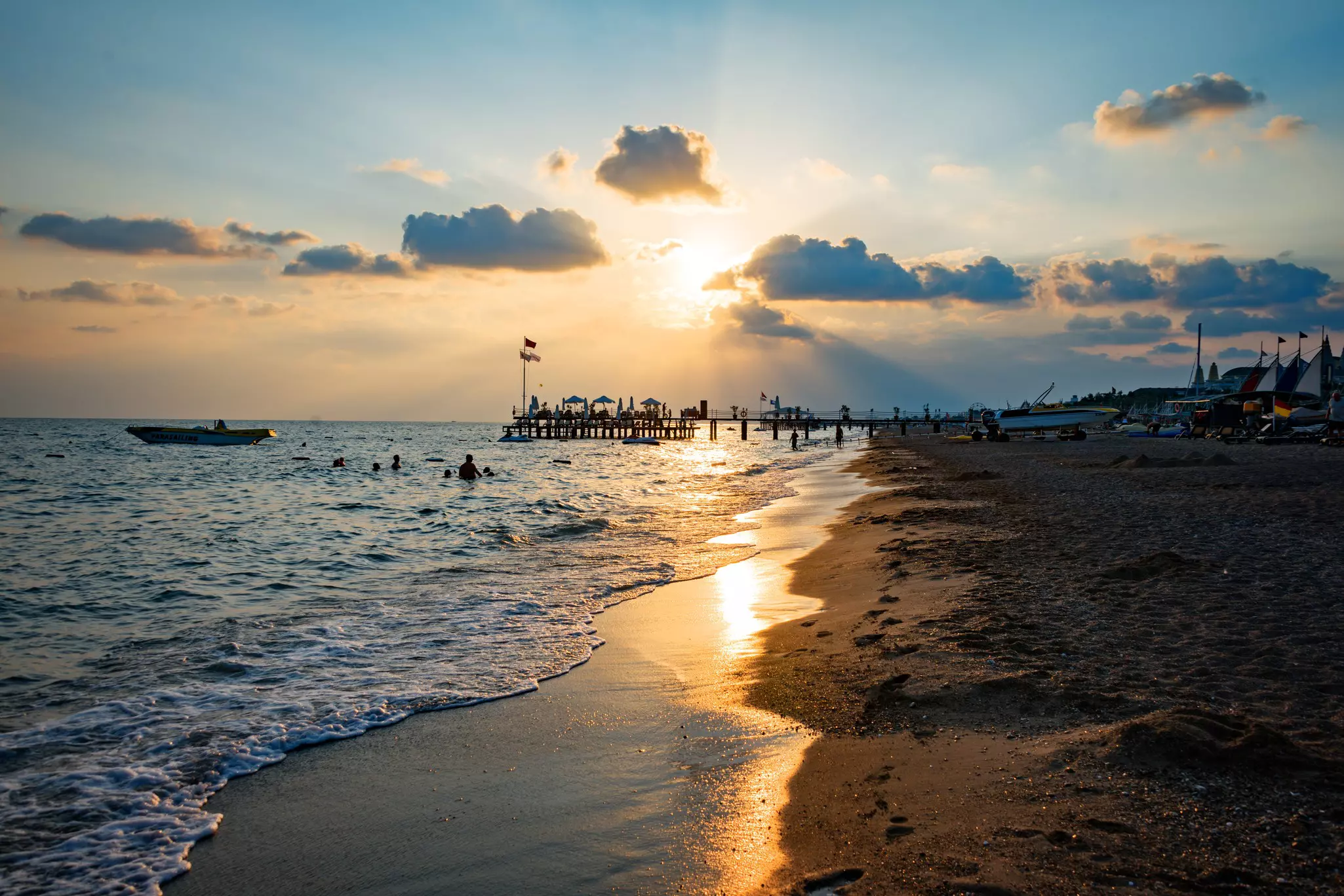 Sunset at Lara Beach, Turkey with a pier in the distance and swimmers in the ocean, a boat floating offshore.
