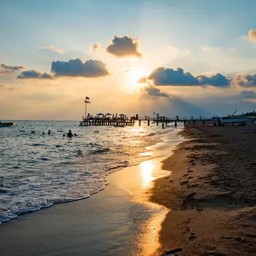 Sunset at Lara Beach, Turkey with a pier in the distance and swimmers in the ocean, a boat floating offshore.