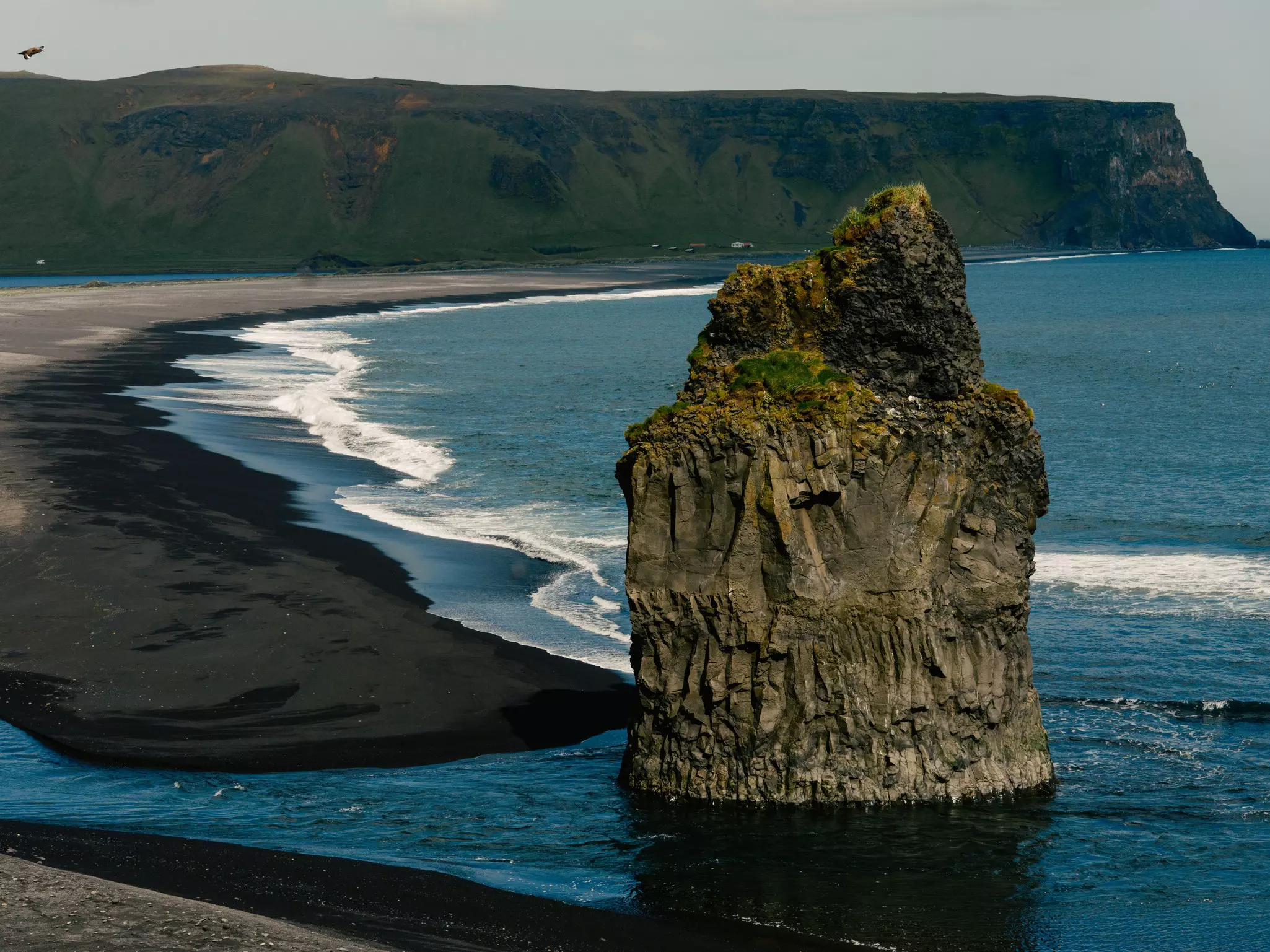 A rock formation in blue water by a black sand beach.