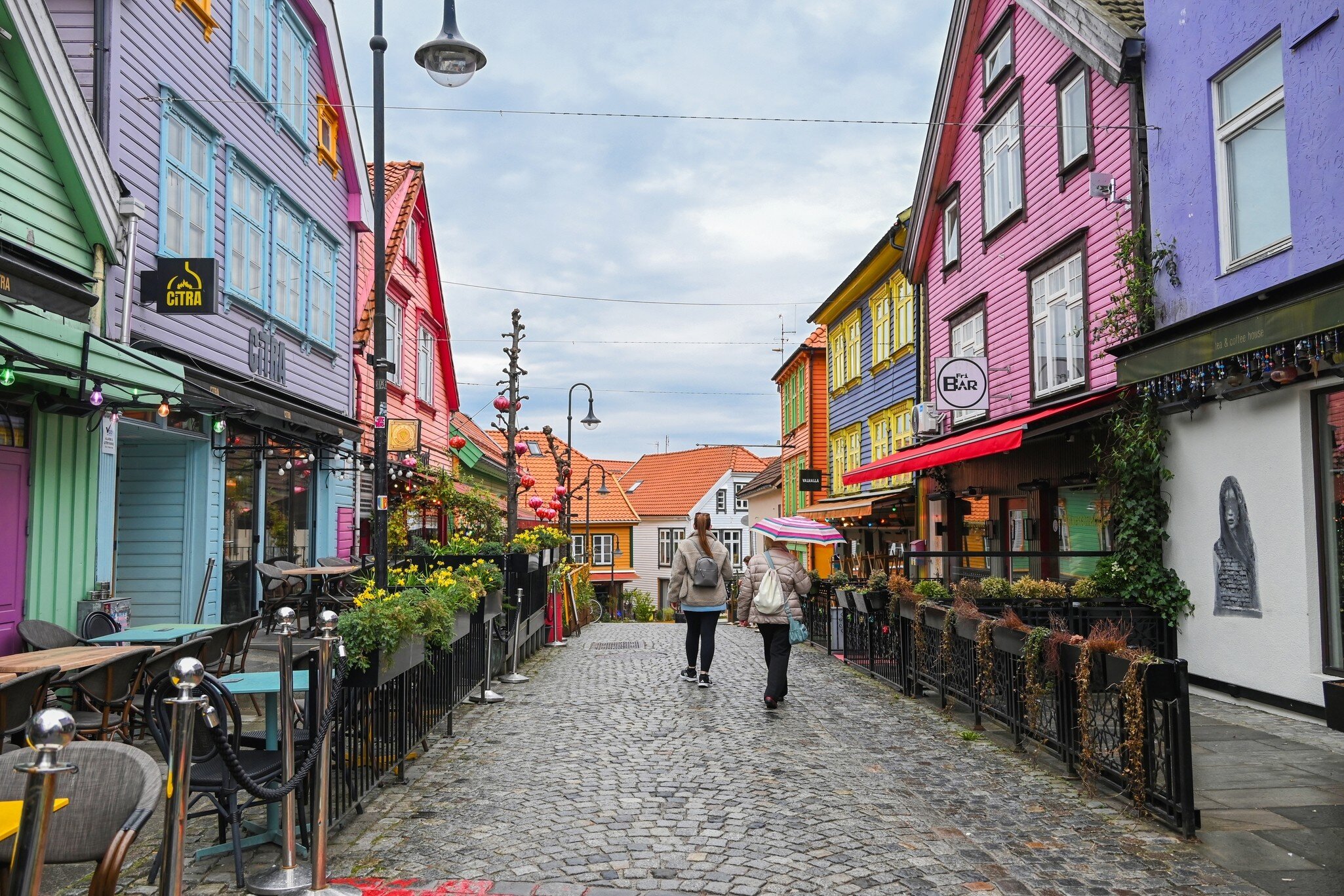 Two people walk a cobbled street lined with bright painted wooden houses in Stavanger, Norway; signs read CITRA and Fri BAR