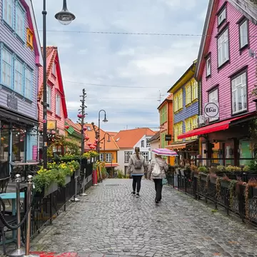 Two people walk a cobbled street lined with bright painted wooden houses in Stavanger, Norway; signs read CITRA and Fri BAR