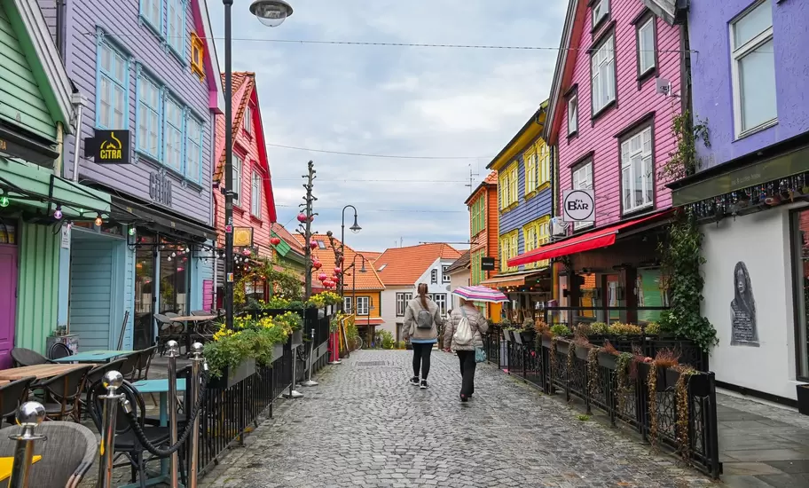 Two people walk a cobbled street lined with bright painted wooden houses in Stavanger, Norway; signs read CITRA and Fri BAR