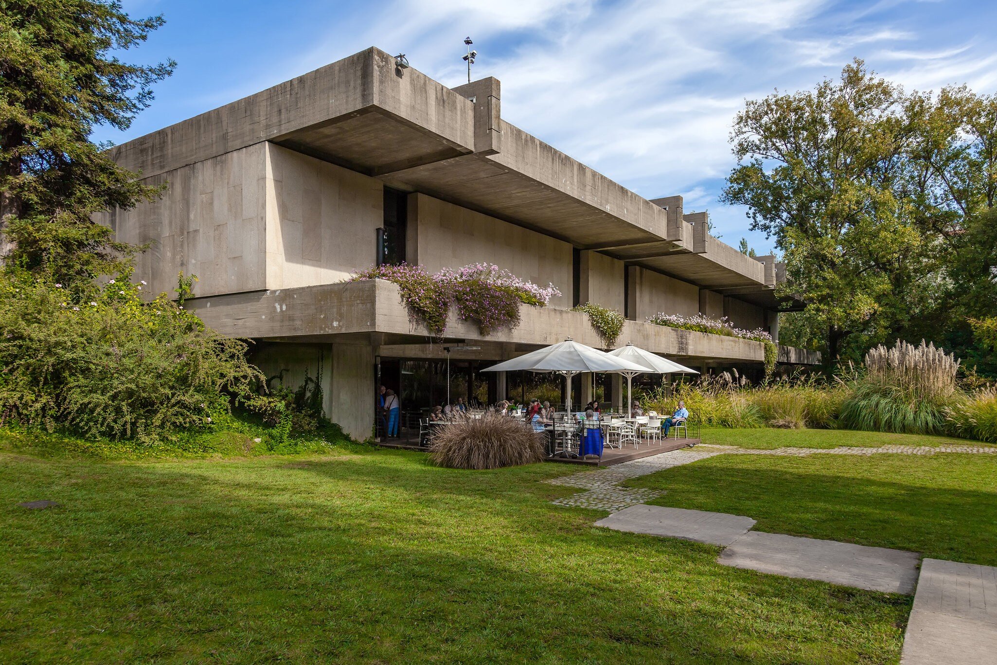 A manicured lawn and large trees surround a Brutalist concrete structure; on the ground level is a cafe with table seating under large umbrellas on a patio.