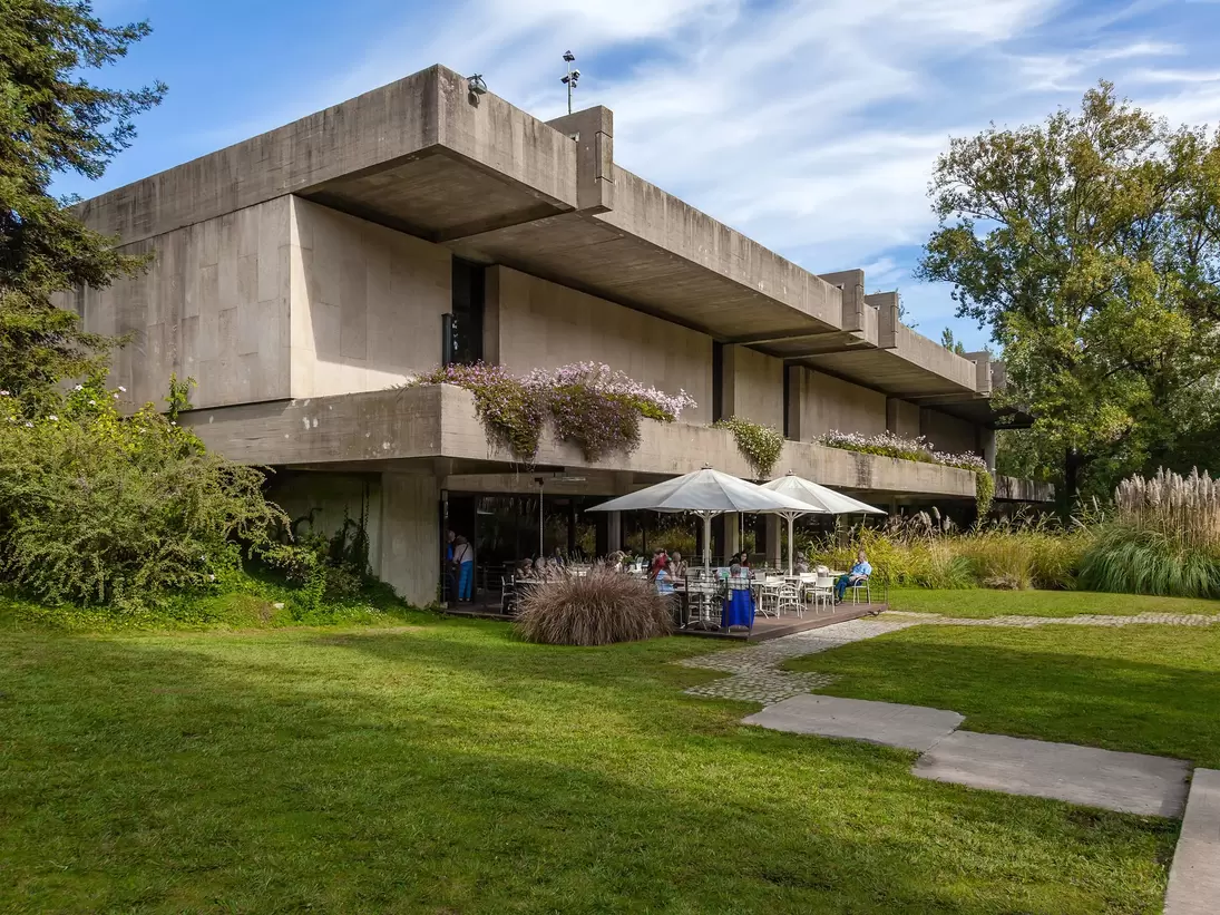 A manicured lawn and large trees surround a Brutalist concrete structure; on the ground level is a cafe with table seating under large umbrellas on a patio.