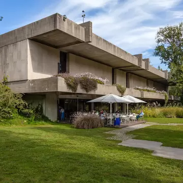 A manicured lawn and large trees surround a Brutalist concrete structure; on the ground level is a cafe with table seating under large umbrellas on a patio.