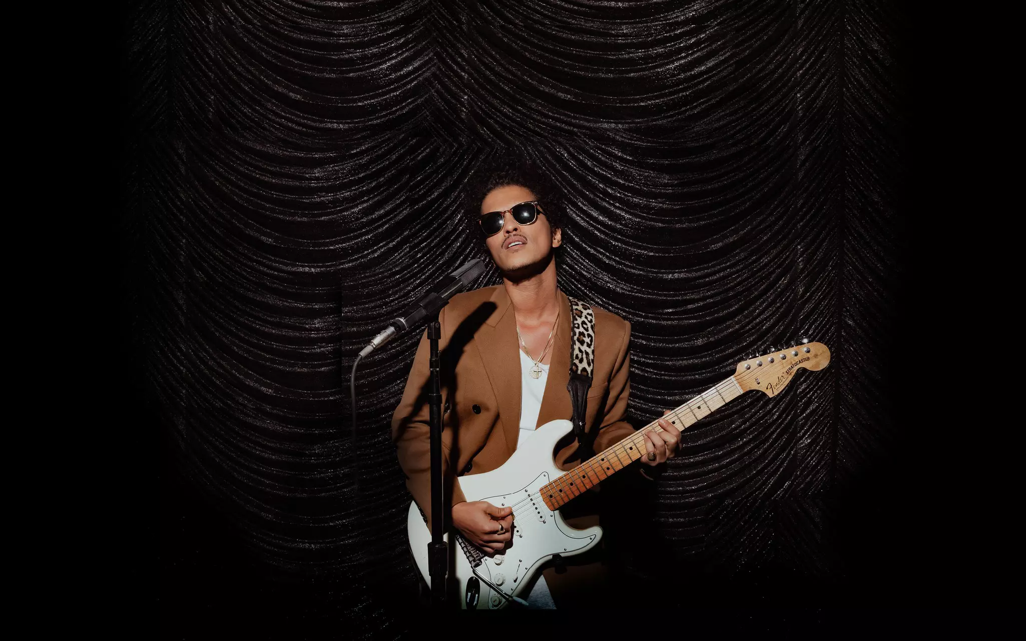 Bruno Mars stands in front of a microphone, holding a cream and white guitar. There's a black curtain in the background.