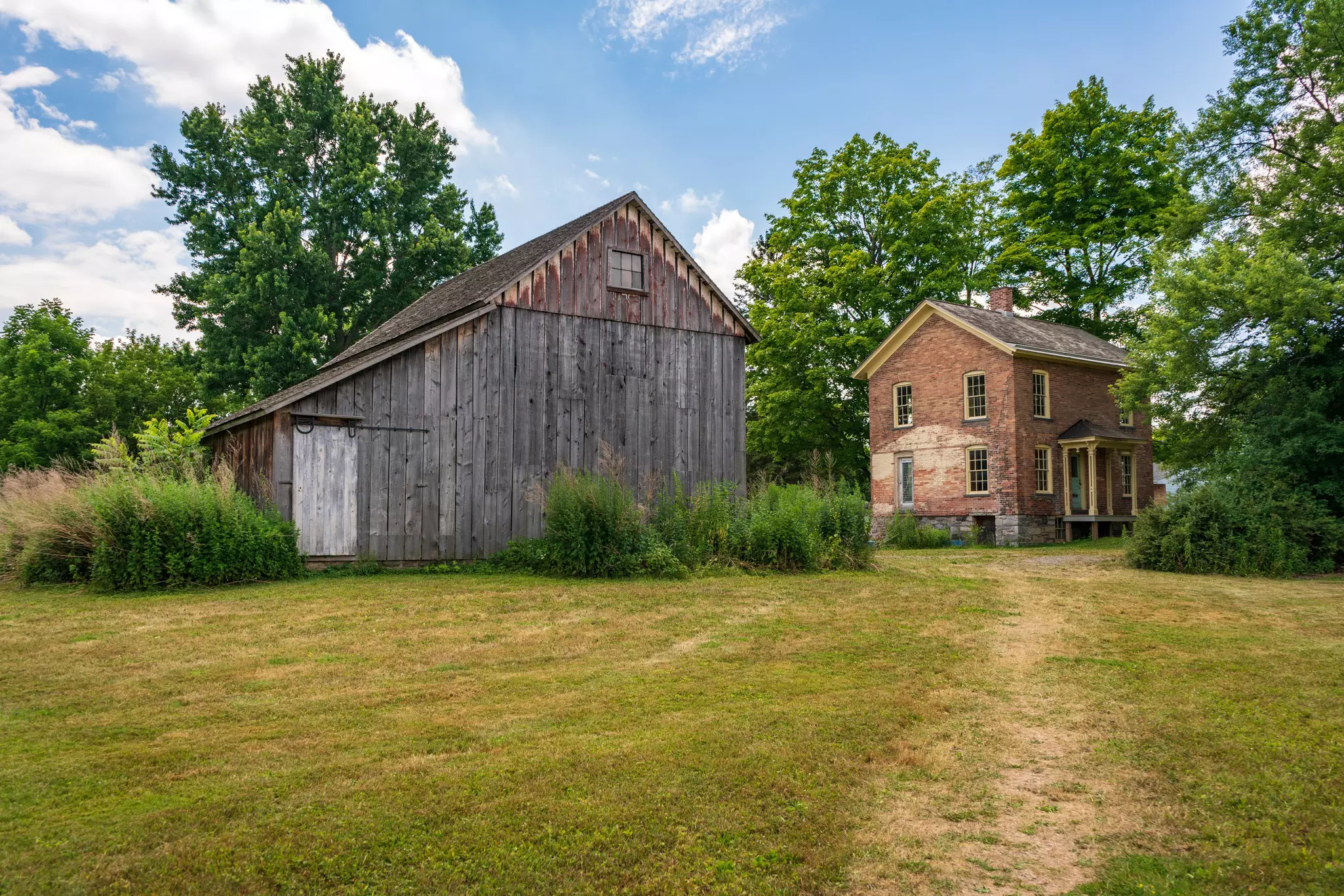 A wooden barn and a two-story red brick house set back in a grass lawn with a worn path to the house.