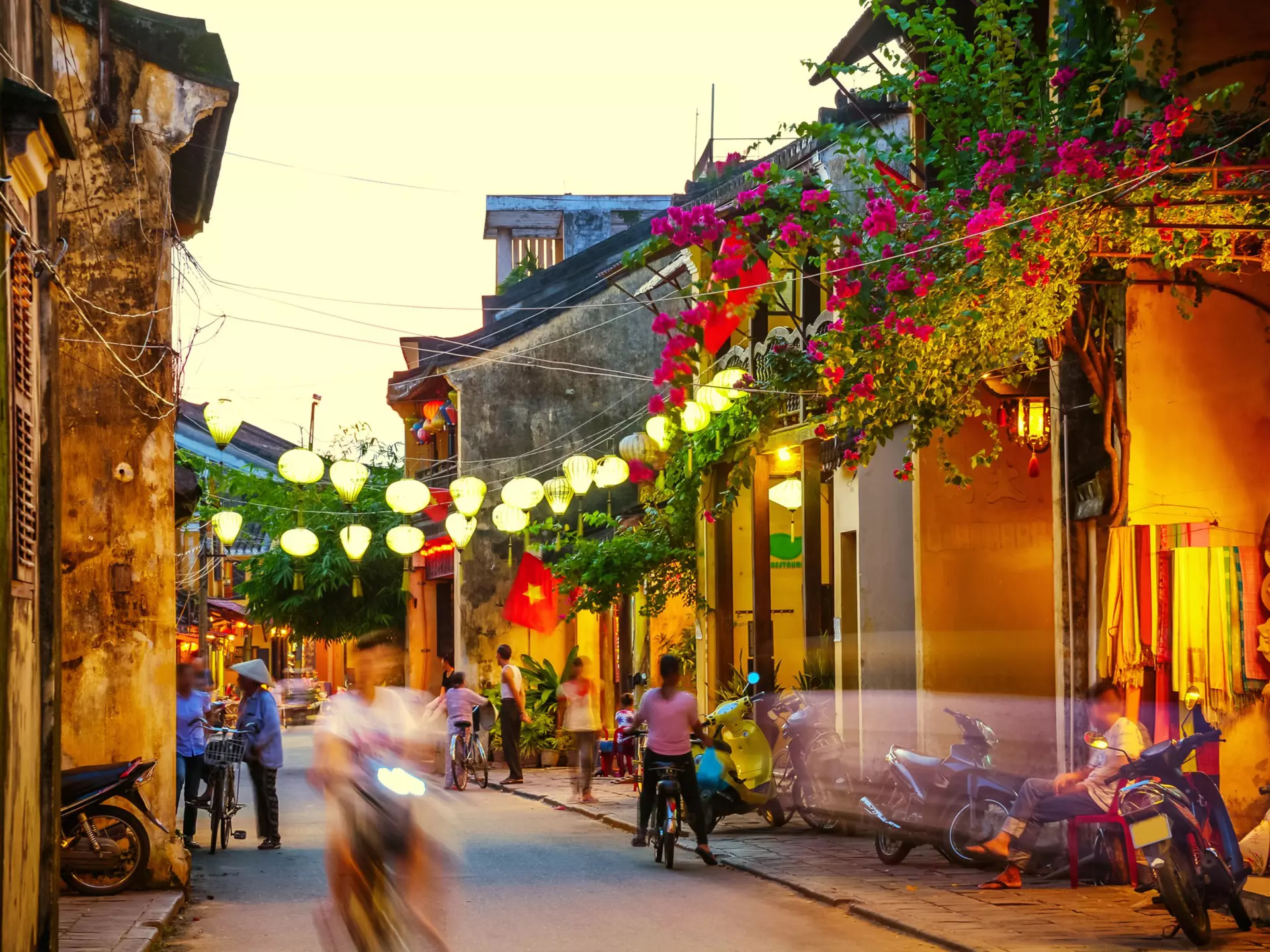 People ride scooters and walk down a narrow street at dusk, lit by lanterns