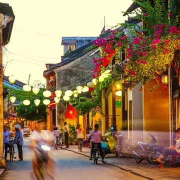 People ride scooters and walk down a narrow street at dusk, lit by lanterns