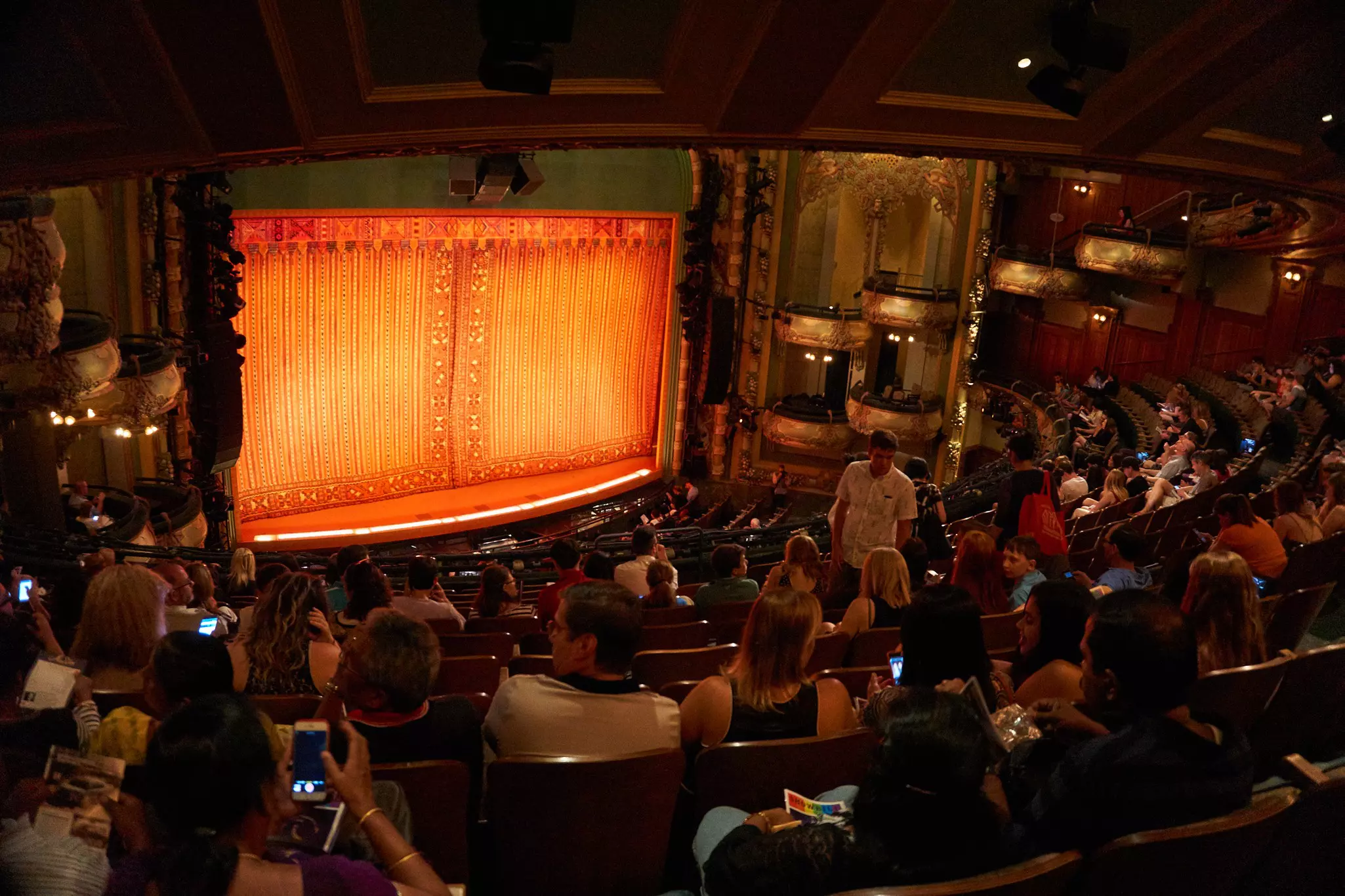 A view of people sitting in rows of seats in a theater. The stage, with a golden curtain, is seen at the front of the house.