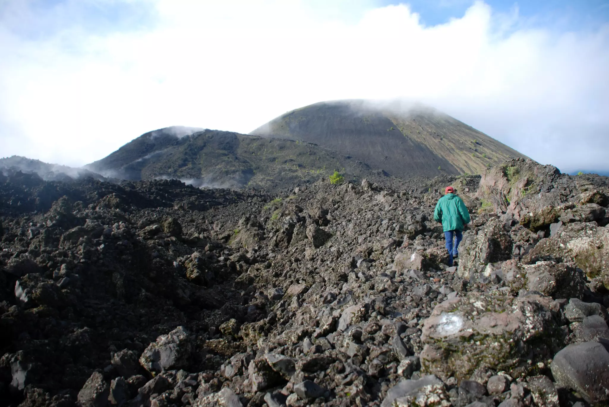 A person in a green park climbs black rocks toward the summit of a volcano.