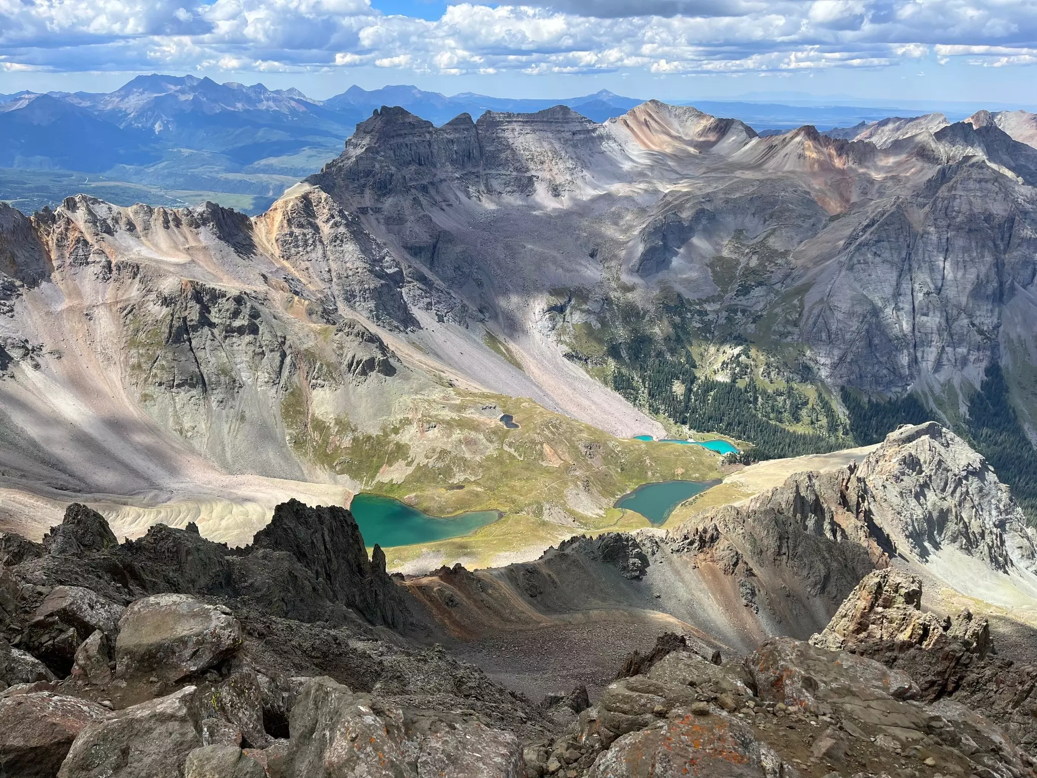 Rugged, rocky mountain range with sandy scree running down to brilliant blue-green lakes on a sunny day with another range of mountains in the distance.
