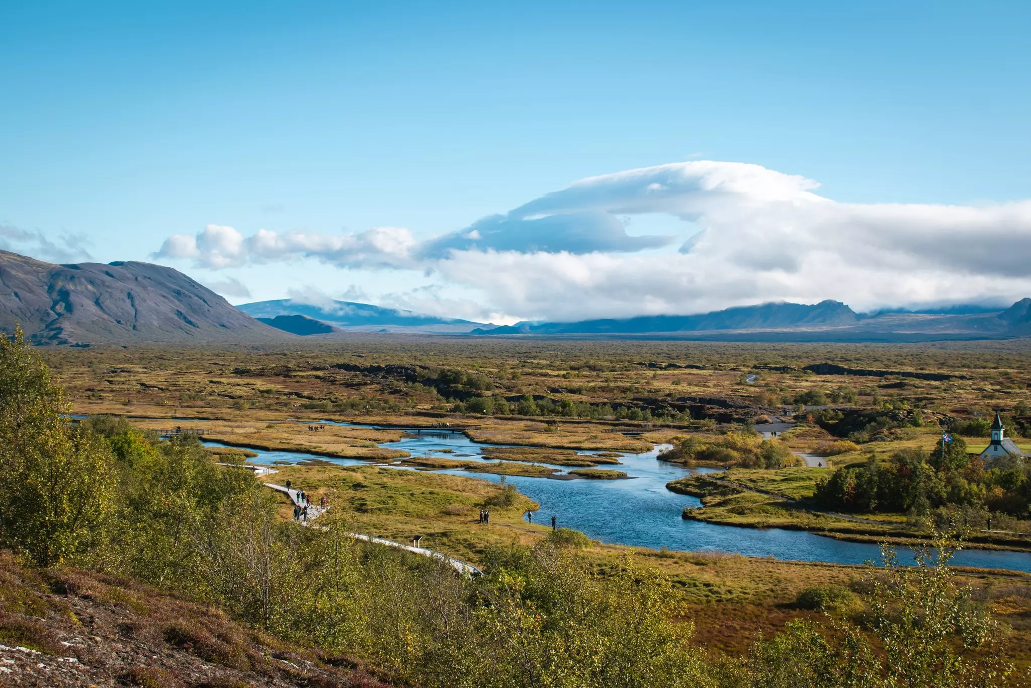 A blue stream in a green landscape with mountains and clouds on the horizon.