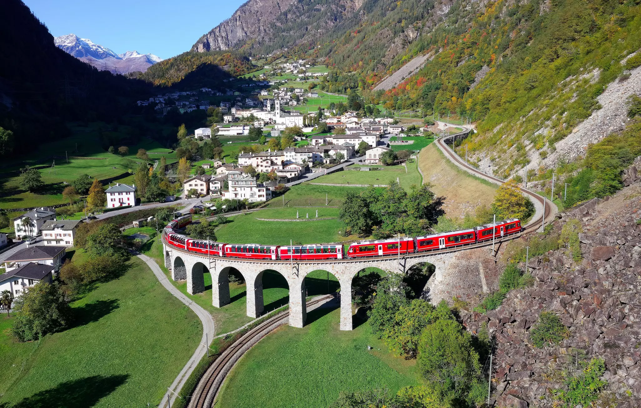 A red train travels down a raised track into a town in a mountain valley.