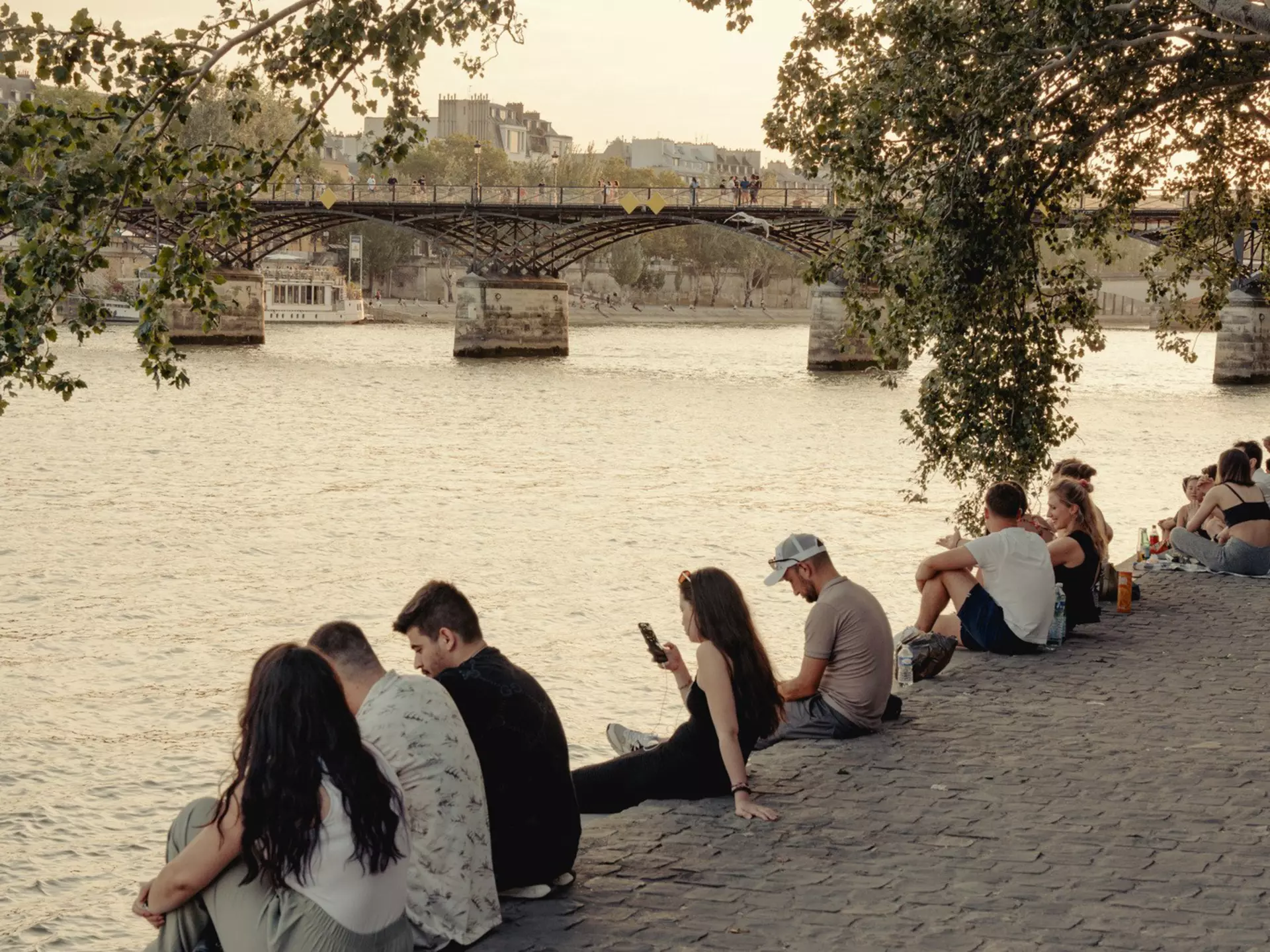 People sit alongside a river in a city during a sunny summer's day