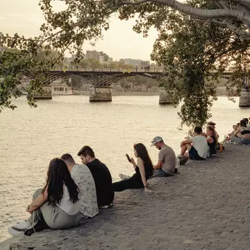 People sit alongside a river in a city during a sunny summer's day
