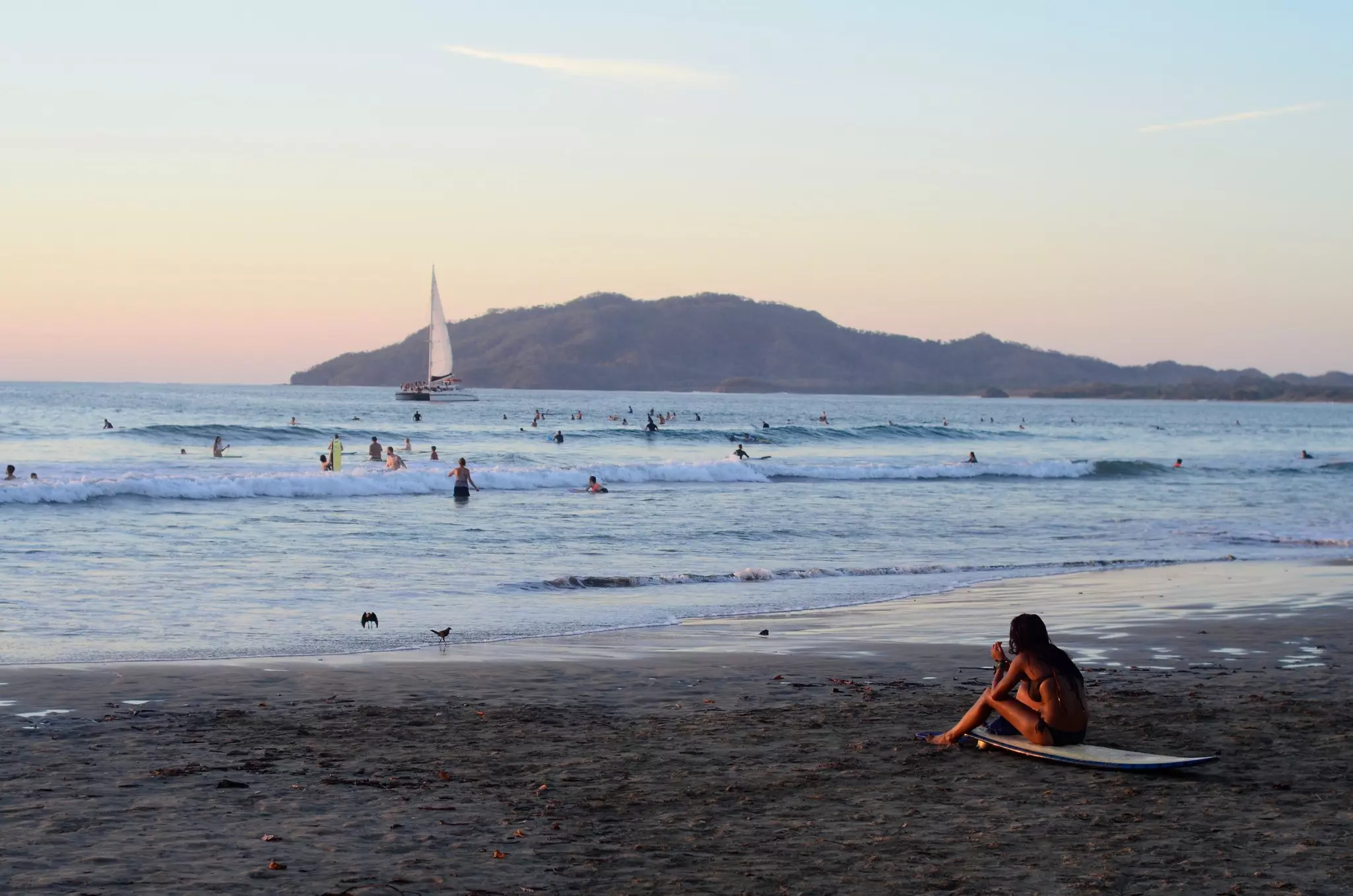 a surfer sits on her board on the beach in Tamarindo, Costa Rica, while people swim and surf in the ocean around sunset