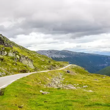 Narrow road curving across a rocky green plateau above a forested valley under heavy cloud on Aurlandsfjellet, Norway