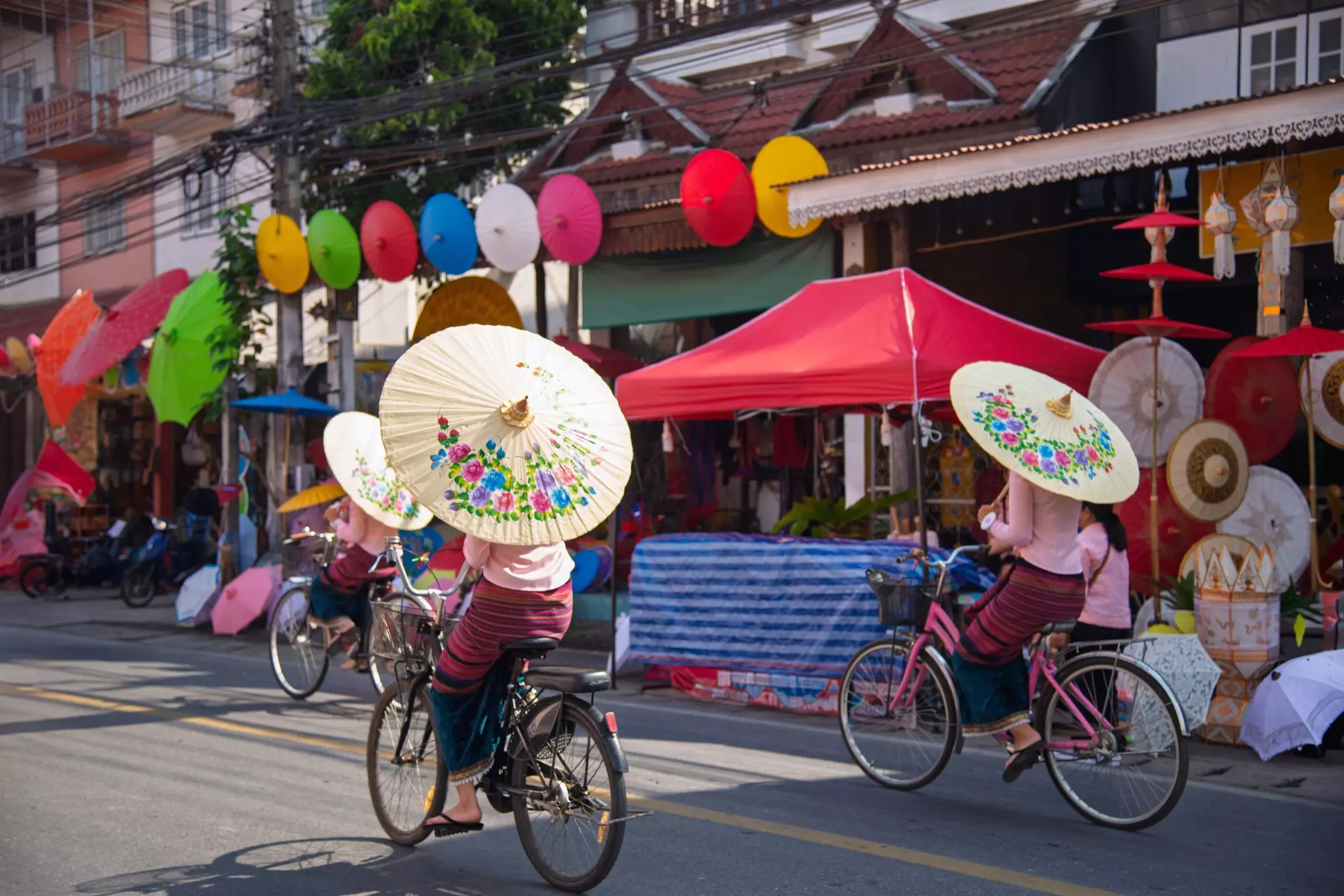 Three women ride bikes on a road while carrying umbrellas decorated with flowers