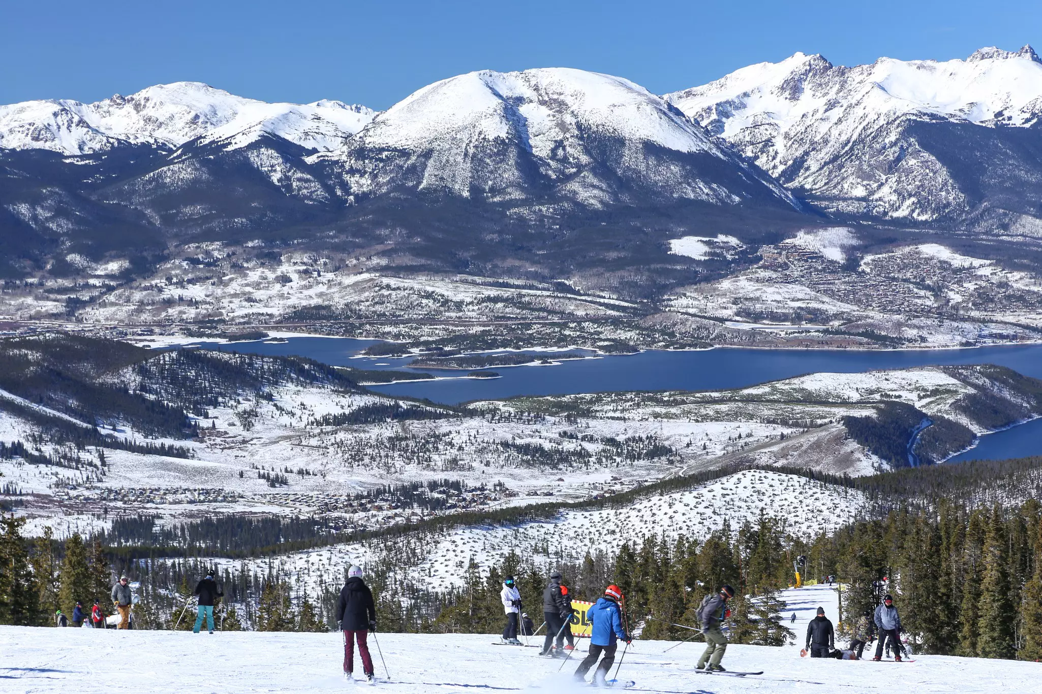 A group of skiers on a mountain above a lake, with more mountains on the other shore.