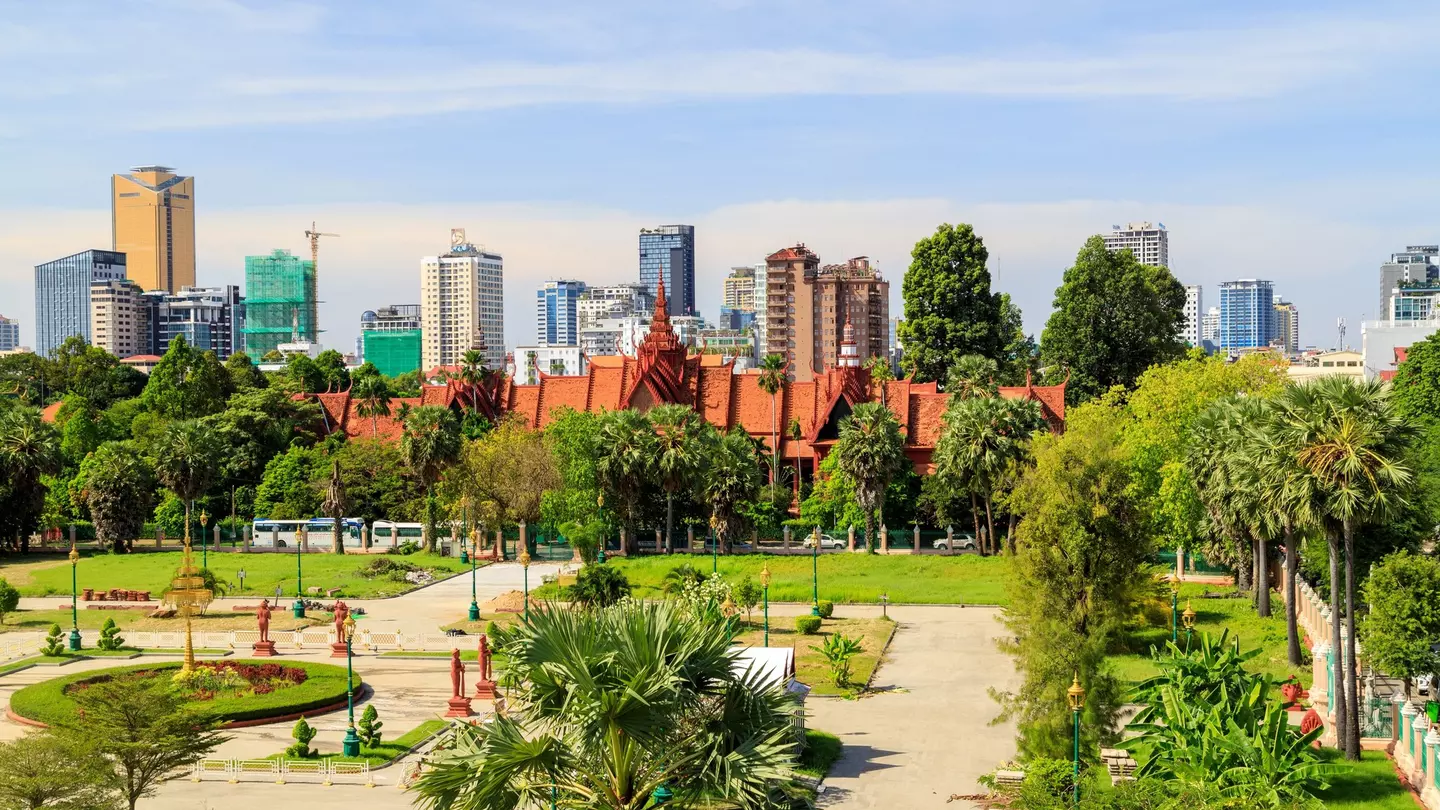 A park in the foreground leads to a building, only visible at its angled red roofline; a city skyline forms the horizon.