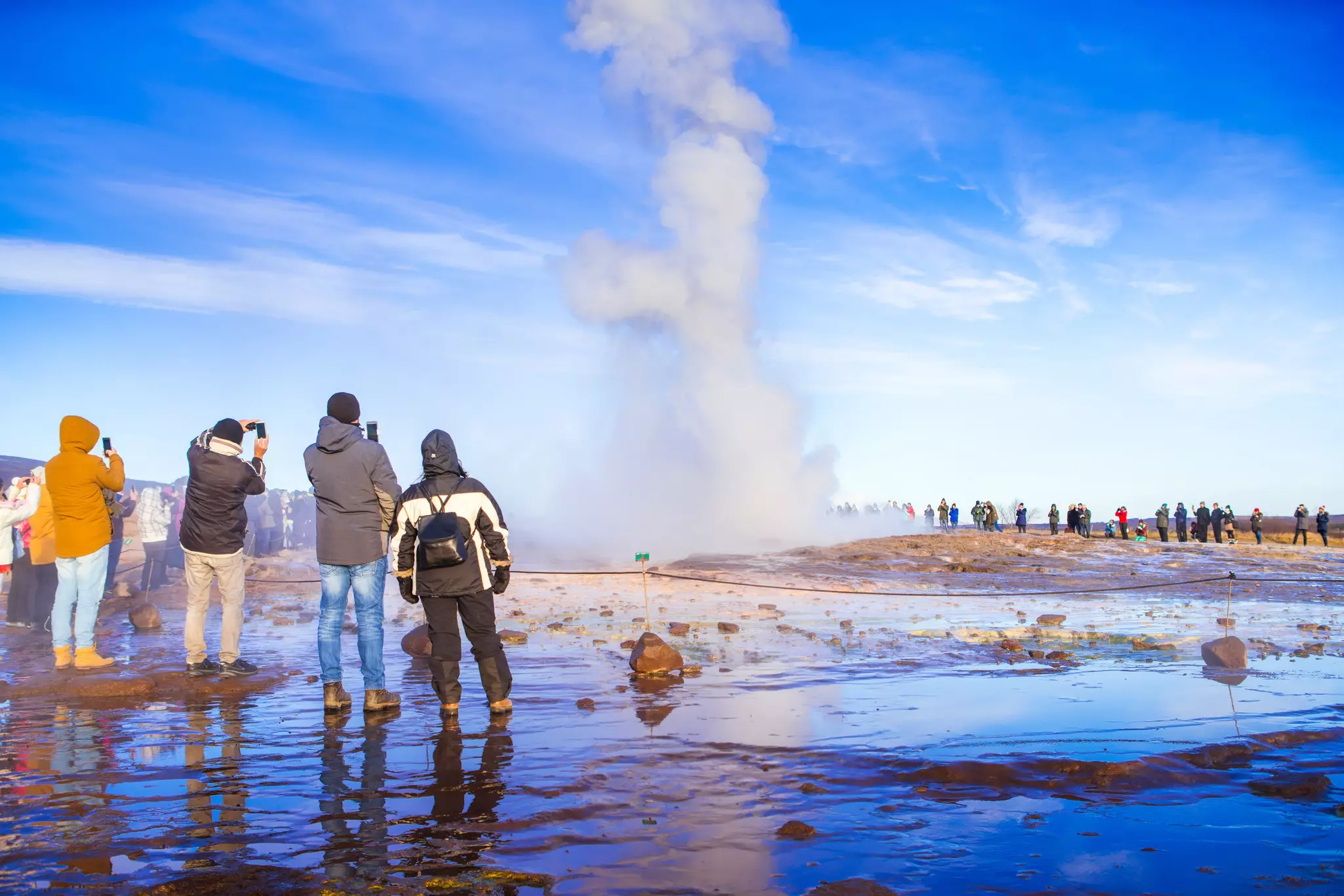People gather at a safe distance to watch an erupting geyser, with water shooting high into the sky.