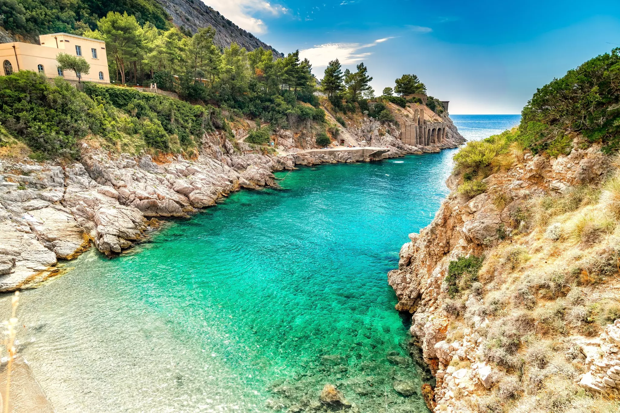 Narrow cove of clear blue-green water surrounded by rocky hills on both sides and a rectangular-roofed house to the left on a sunny day.