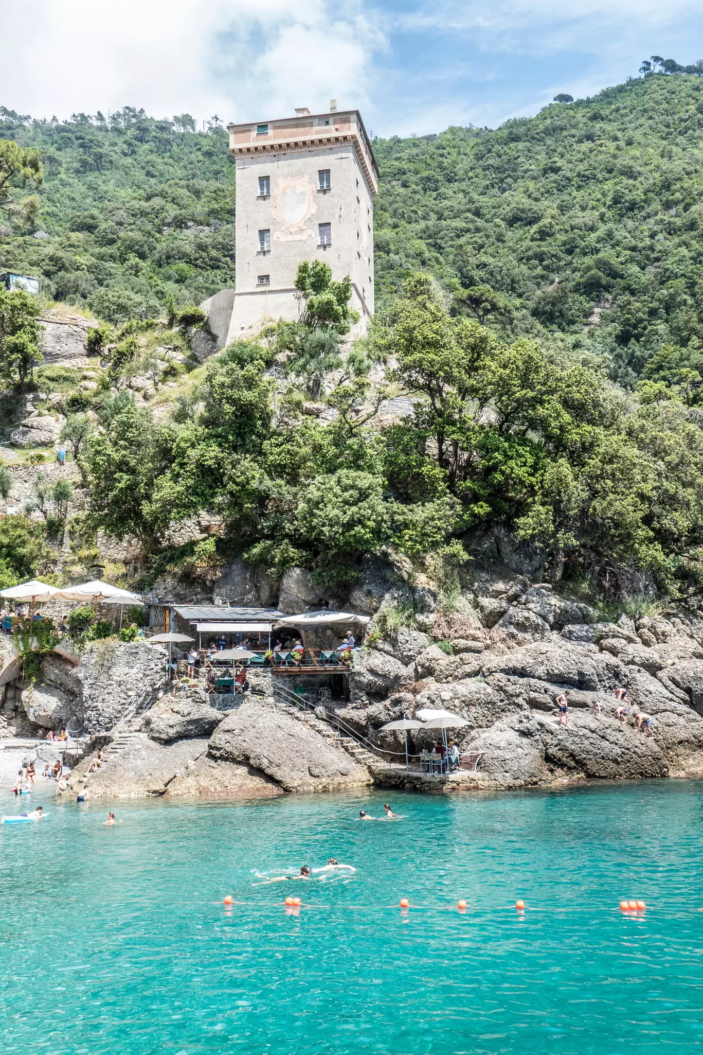 People swim in blue/green water with small tables on a cliffside in the background. Above is the tower of an abbey.