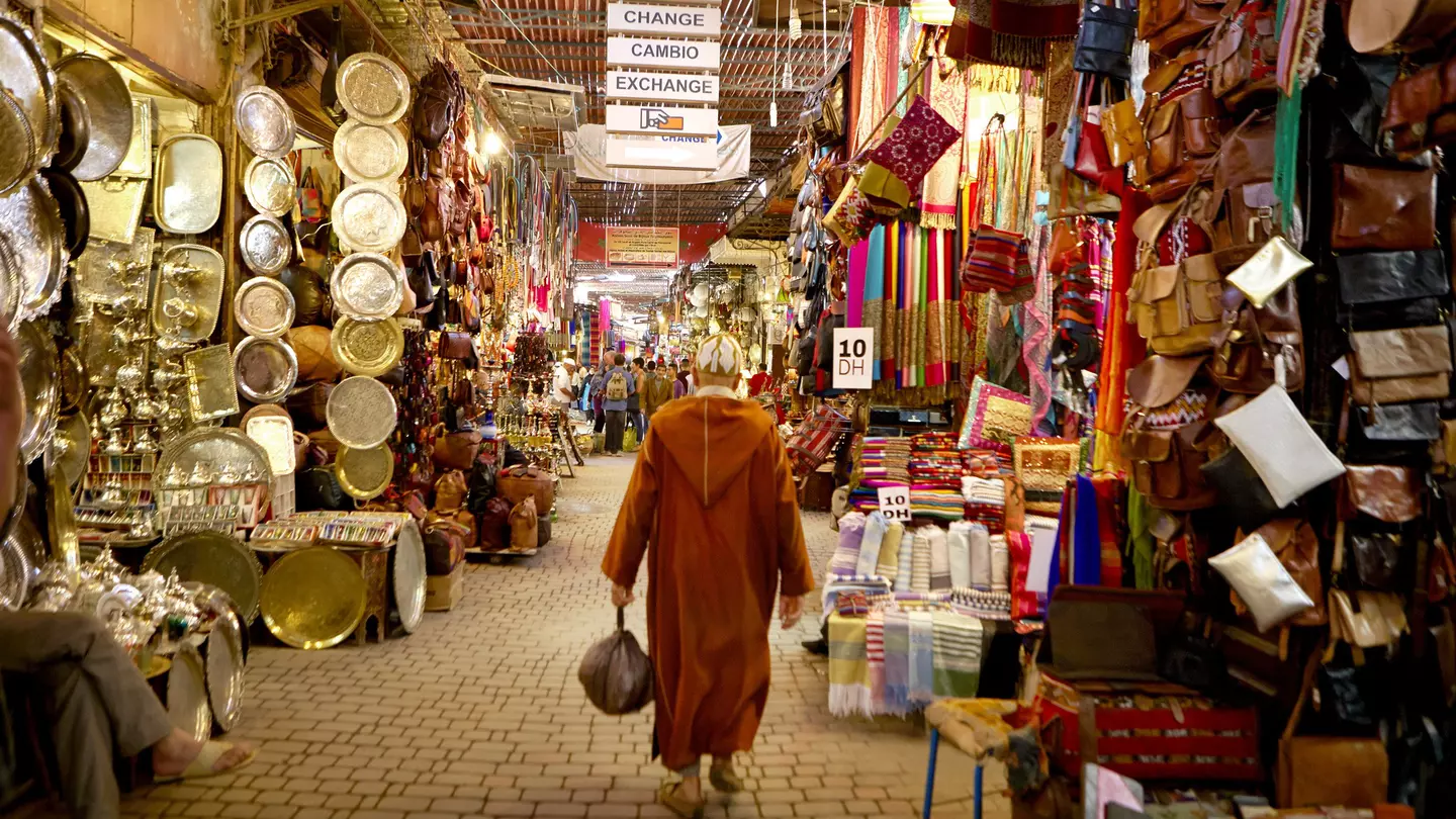 A man in traditional Moroccan robe called a Djellabah walks away from the camera. Stalls selling plates, bags and scarves line either side.