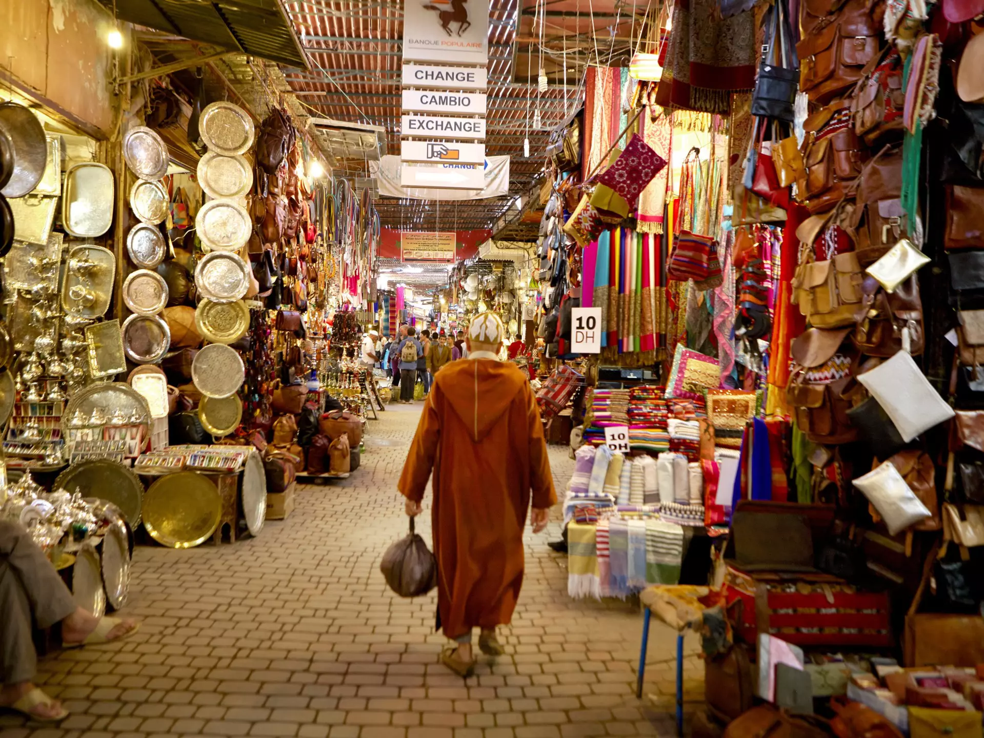 A man in traditional Moroccan robe called a Djellabah walks away from the camera. Stalls selling plates, bags and scarves line either side.