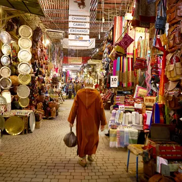 A man in traditional Moroccan robe called a Djellabah walks away from the camera. Stalls selling plates, bags and scarves line either side.
