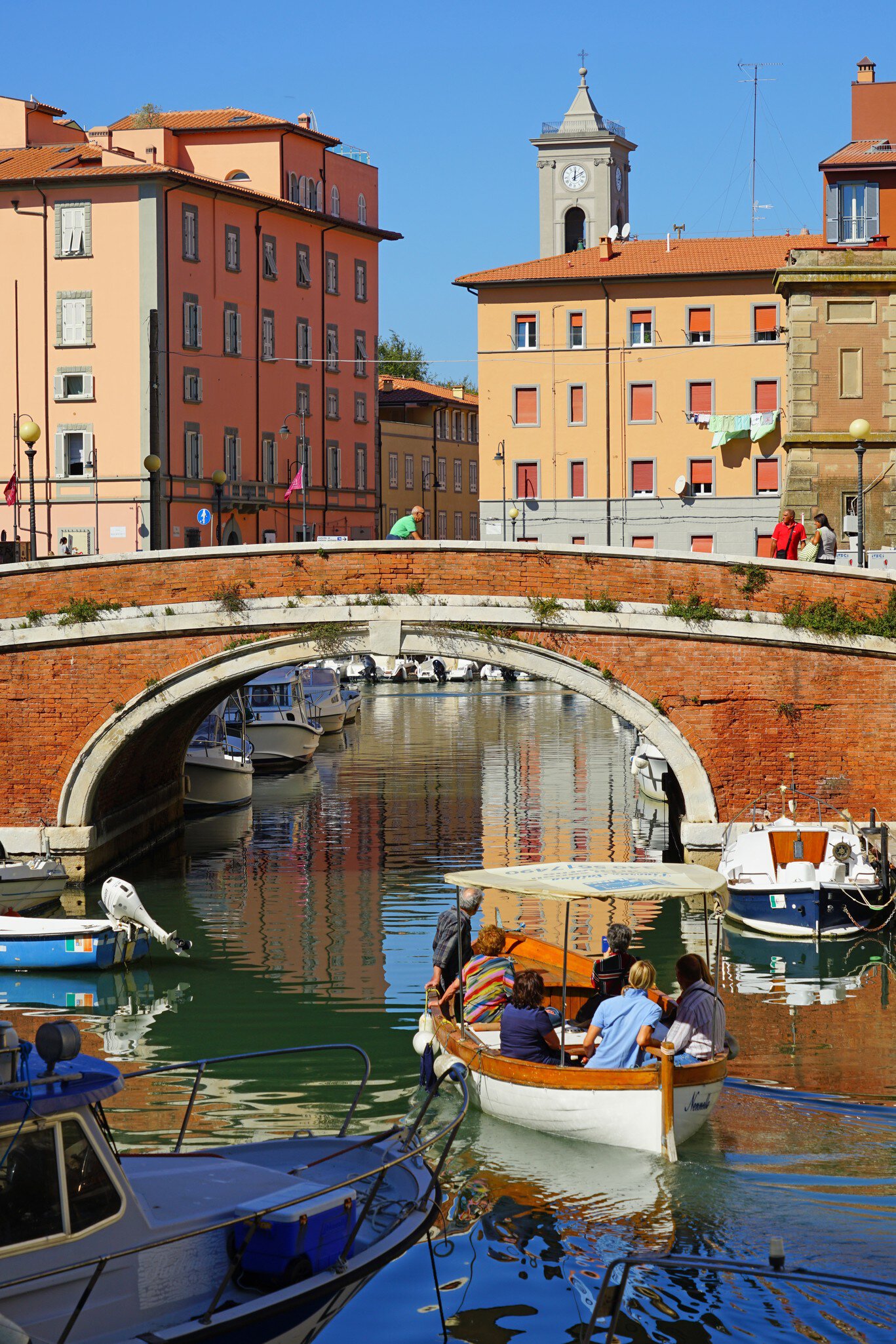 A canopied wooden launch with six passengers on a Livorno canal, past moored motorboats, pastel houses and a clock tower.