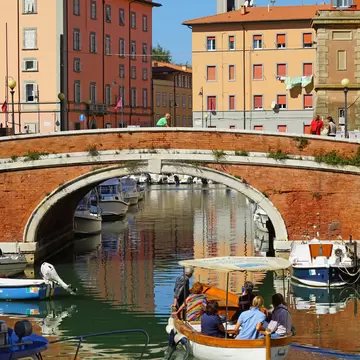 A canopied wooden launch with six passengers on a Livorno canal, past moored motorboats, pastel houses and a clock tower.