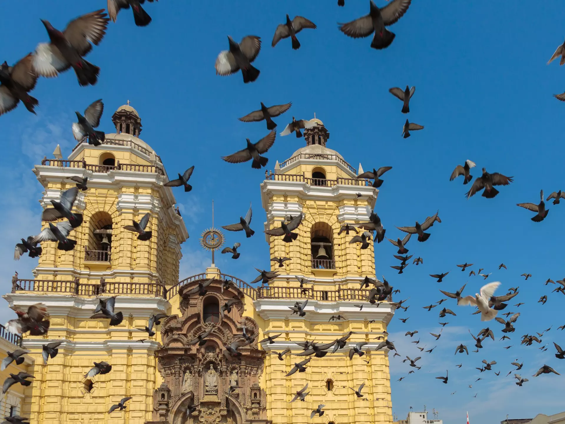 A flock of pigeons fly upwards into a blue sky above a yellow church building with two prominent bell towers