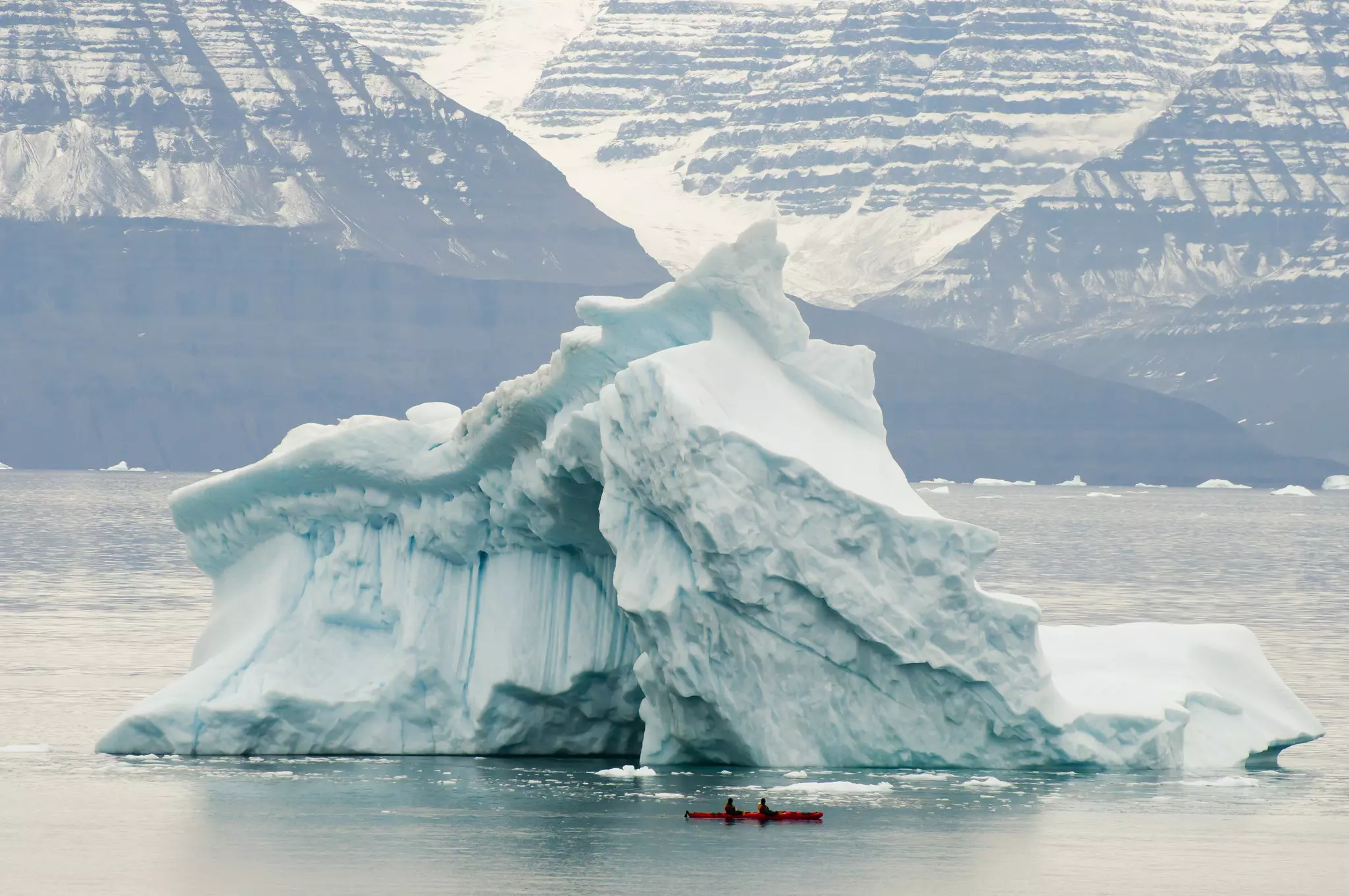 Two people in a tandem kayak float past the base of a large iceberg in Greenland.