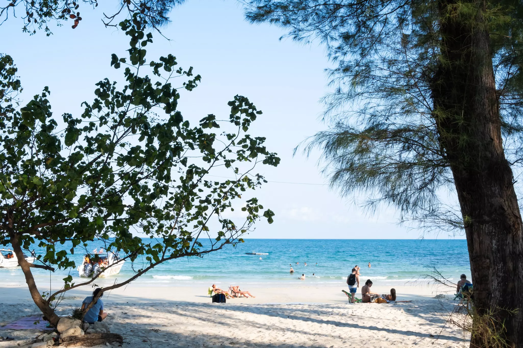 Sunbathers on a white-sand beach with some trees casting shade