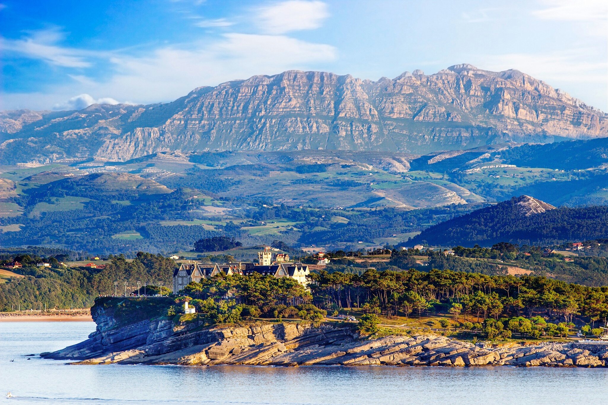 Turreted palace among pine trees on a rocky headland above calm water, with layered green hills and a limestone ridge beyond.