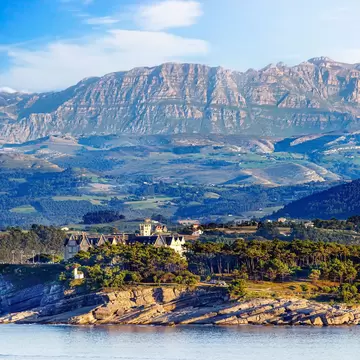 Turreted palace among pine trees on a rocky headland above calm water, with layered green hills and a limestone ridge beyond.