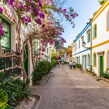 White buildings with brightly colored trip on a pedestrian street; there is a pink blooming vine growing on one in the foreground.