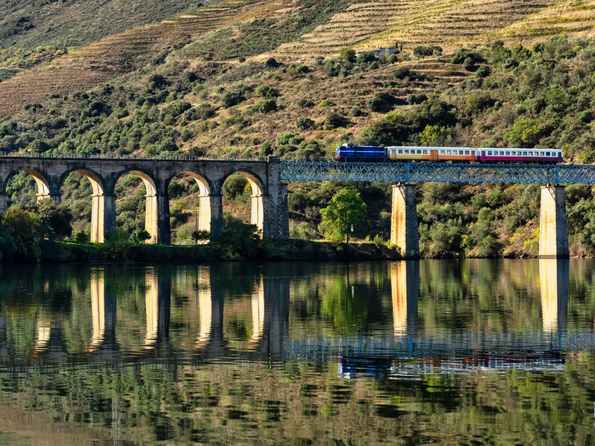 A train on a bridge crossing a river, with the hillside reflected in the water's surface.