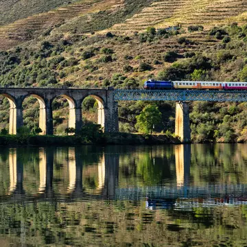 A train on a bridge crossing a river, with the hillside reflected in the water's surface.