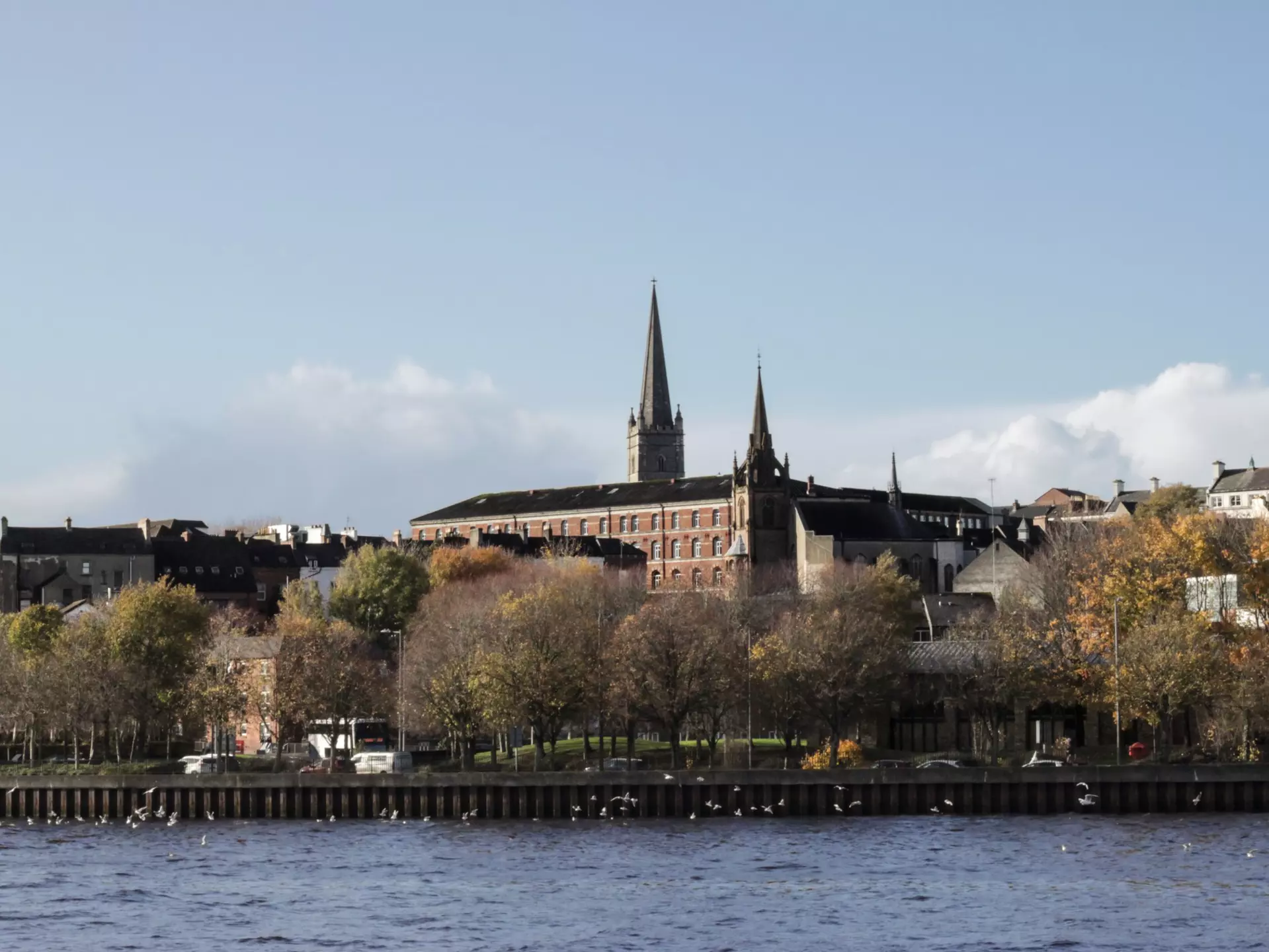 A city on a riverbank, with two spires rising above the trees and buildings.