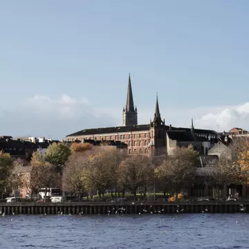 A city on a riverbank, with two spires rising above the trees and buildings.