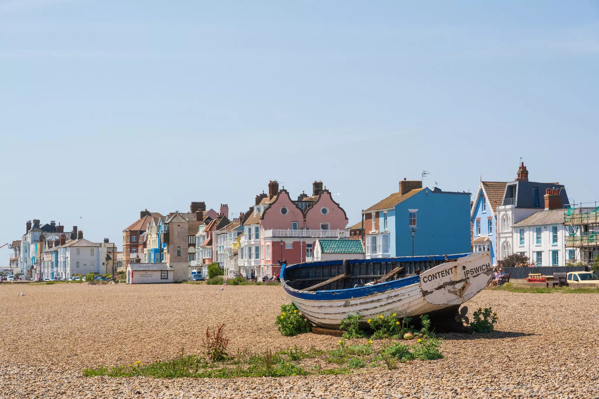 An old wooden fishing boat leans on its side on a a pebbly beach near pastel-painted houses of the town.