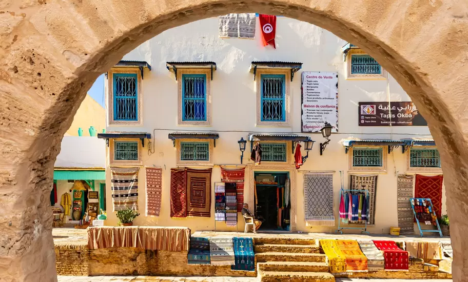 Rugs hung outside the Tapis Okba carpet shop in Tunisia, seen through a sandstone arch, Tunisian flag above