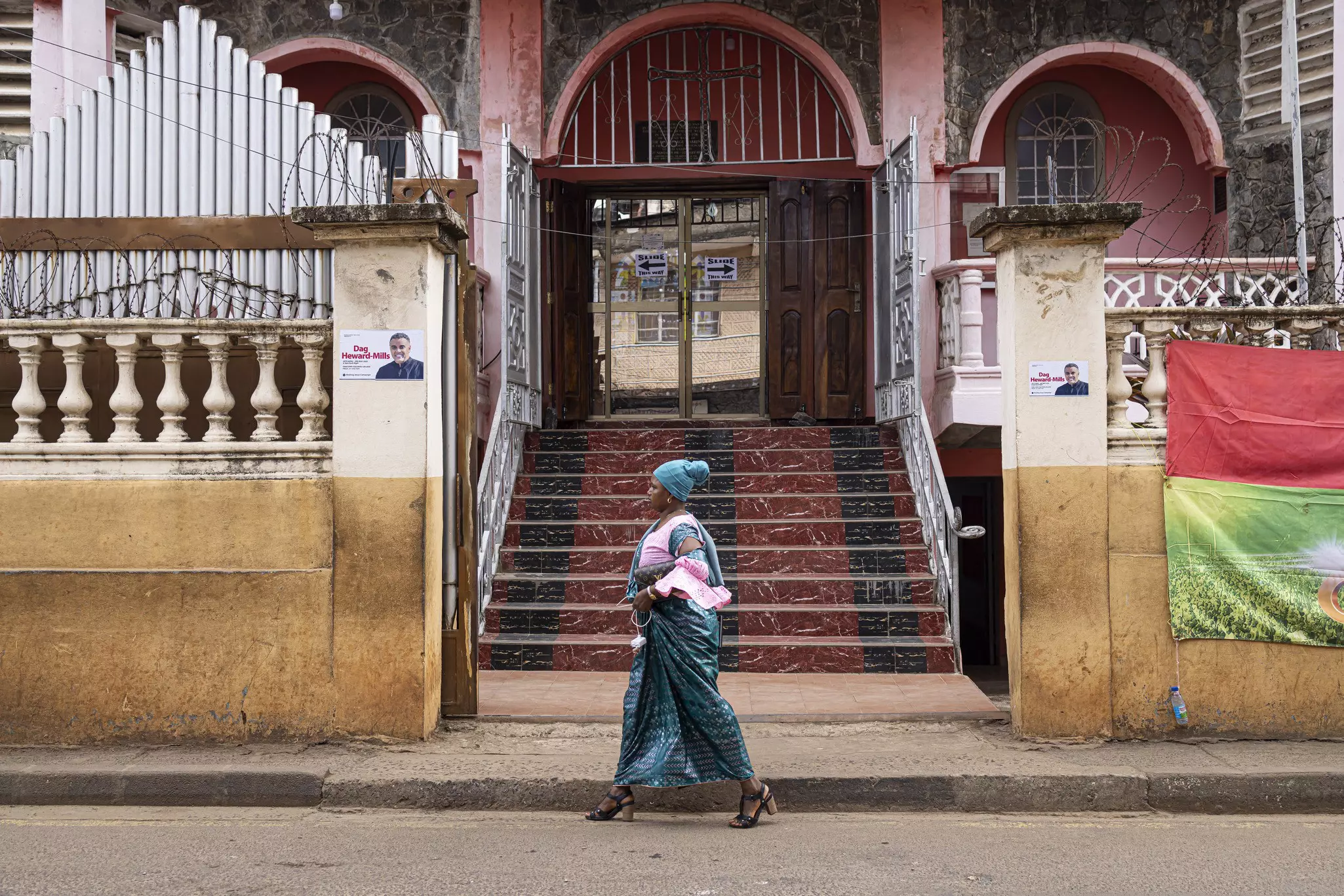 A woman in a green dress and hat walks on a street in a city past stairs leading to a church.