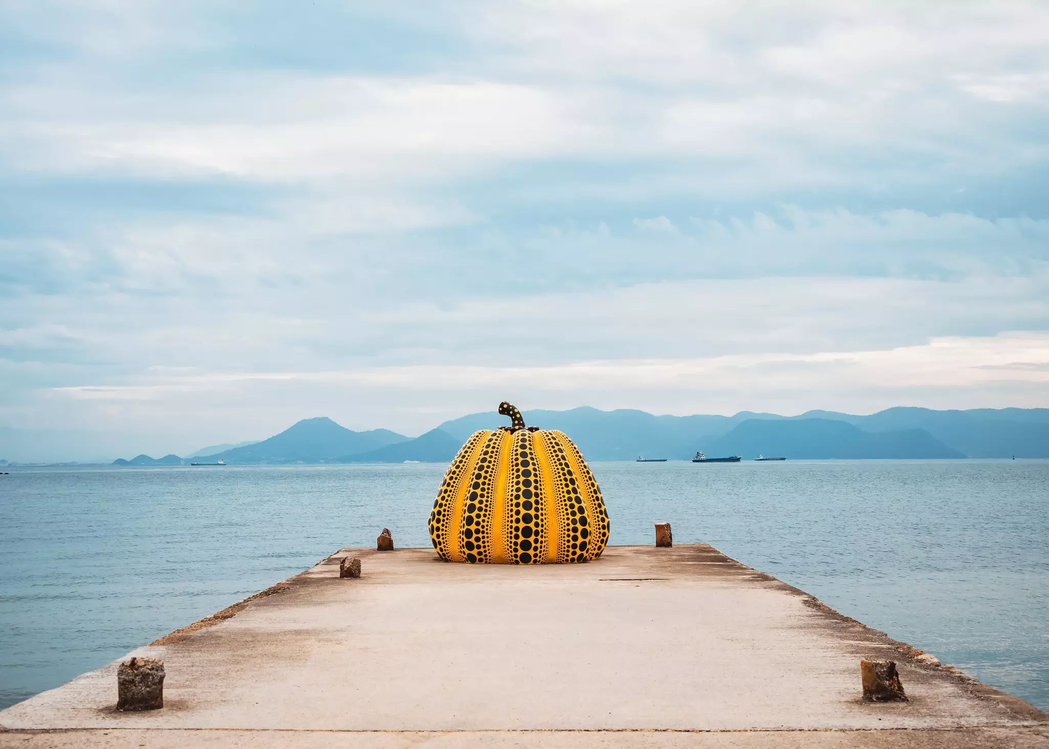 Sculpture of a yellow pumpkin with black dots on a dock overlooking water, with mountains in the distance