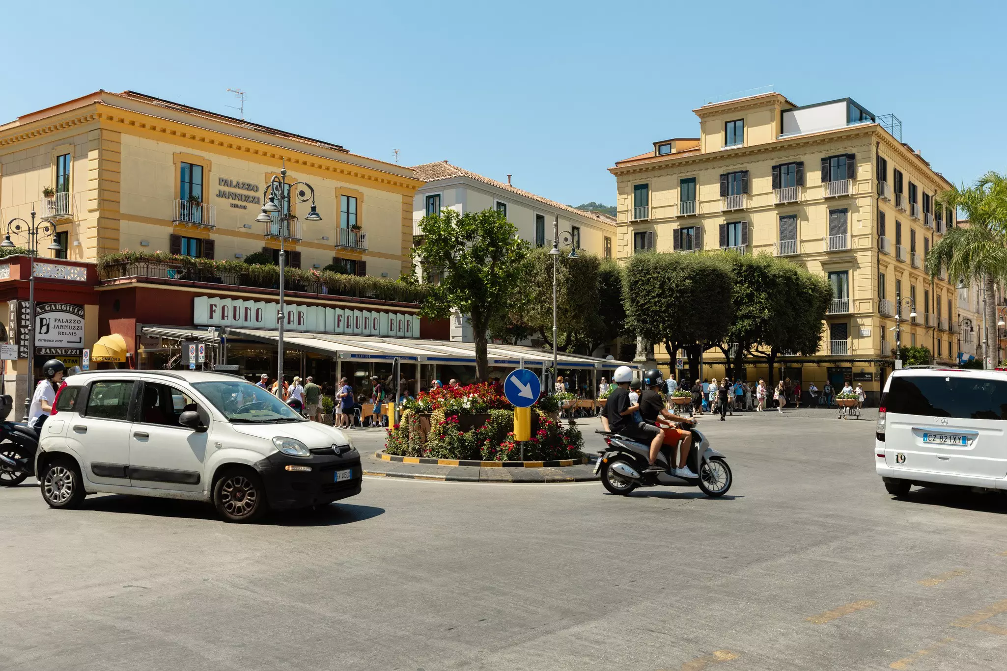 A car, Vespa and minivan make their way through a street lined with yellow and white low-profile buildings with many windows