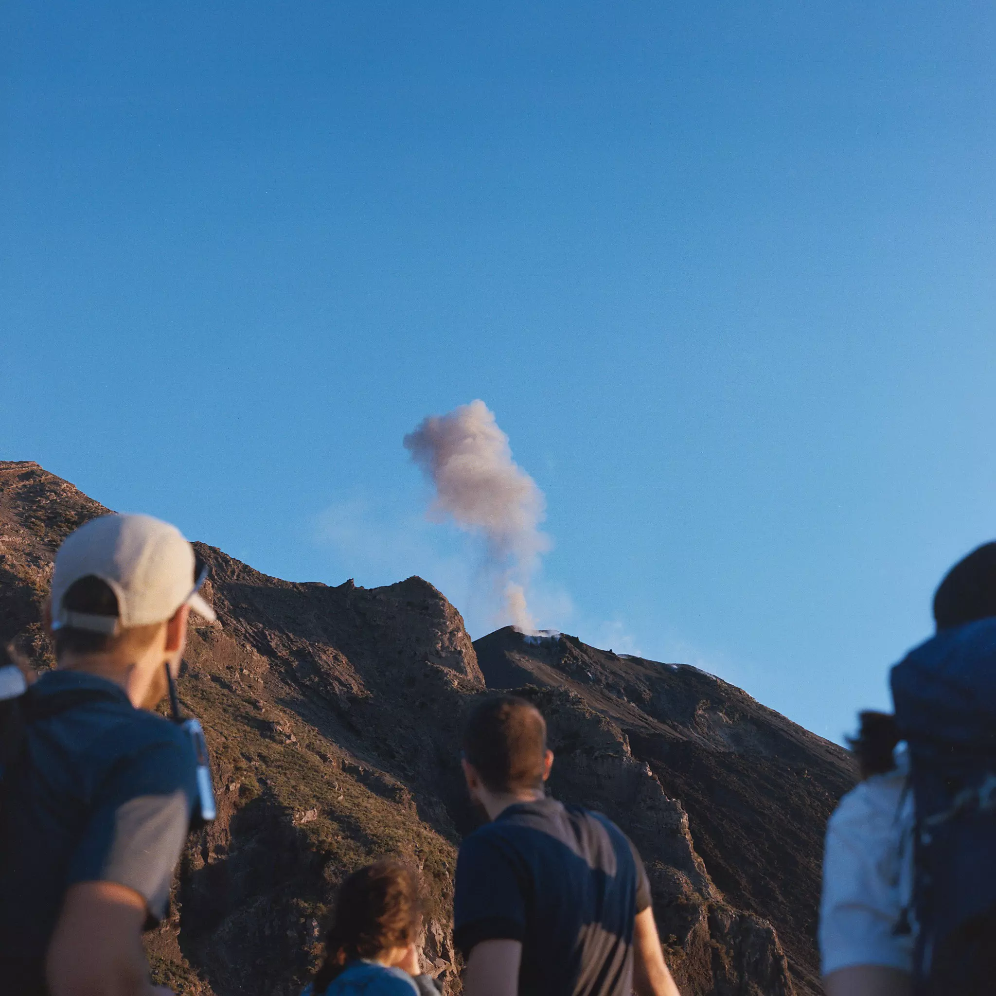 A group of young people look up a rocky slope to see a plume of smoke emerging from a volcano.